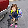 child, girl, smiling, backpack, denim_jacket, floral_dress, pink_clogs, road, sidewalk, vehicle, tire, grass, outdoor, blue_eyes, long_hair, colorful, casual_clothing, happy, standing, daytime