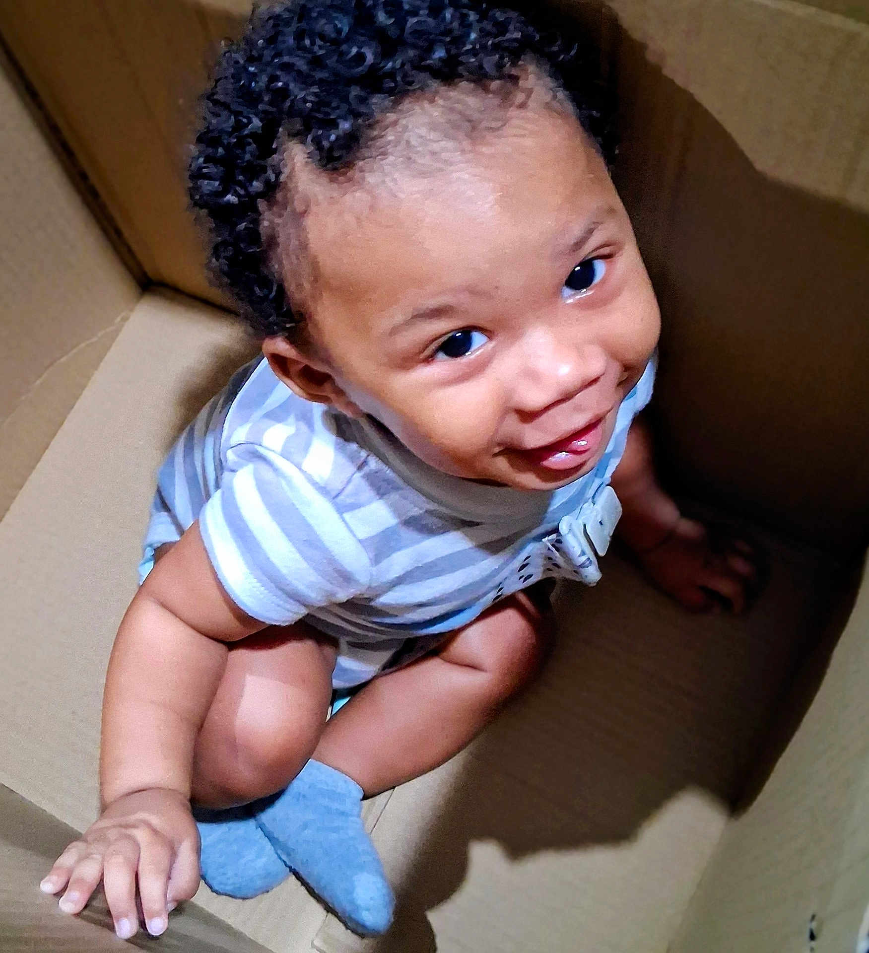Amri is registered to the contest to win money with this photo: baby, infant, toddler, child, smiling, curly_hair, onesie, striped_clothes, cardboard_box, sitting, looking_up, big_eyes, socks, hand, cute, playful, indoor, shadow, closeup, portrait
