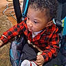 baby, black_pants, buckle, car_seat, child, curly_hair, cushion, cute, hand, infant, looking_sideways, pacifier_clip, plaid, portrait, red_plaid_shirt, seat_padding, sock, teether, toy, wooden_floor