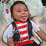 Amri is registered to the contest to win money with this photo: baby, child, infant, portrait, smiling, tongue_out, curly_hair, sitting, costume, pirate, striped_shirt, skull_and_crossbones, red, black, white, cushion, blanket, chair, indoor, happy