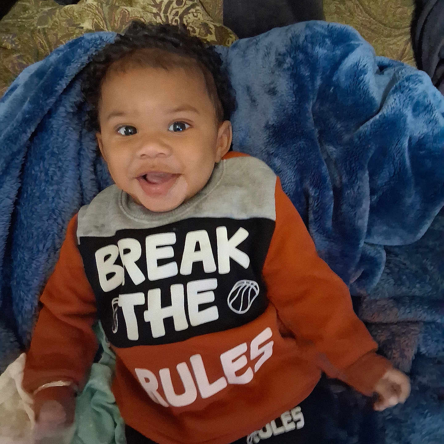 Amri joined the competition — help win amazing prizes! baby, child, infant, smiling, happy, portrait, sitting, face, eyes, curly_hair, orange_clothing, sweatshirt, text_on_clothing, blanket, blue_blanket, indoor, couch, cozy, playful, photography