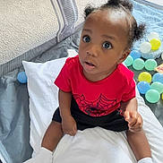 Omari is registered to the contest to win money with this photo: toddler, child, baby, pillow, playpen, plastic_balls, red_shirt, spider_web, socks, carpet, indoor, curious, sitting, person, cute, hair_puffs, toy, floor, looking_up, young_child