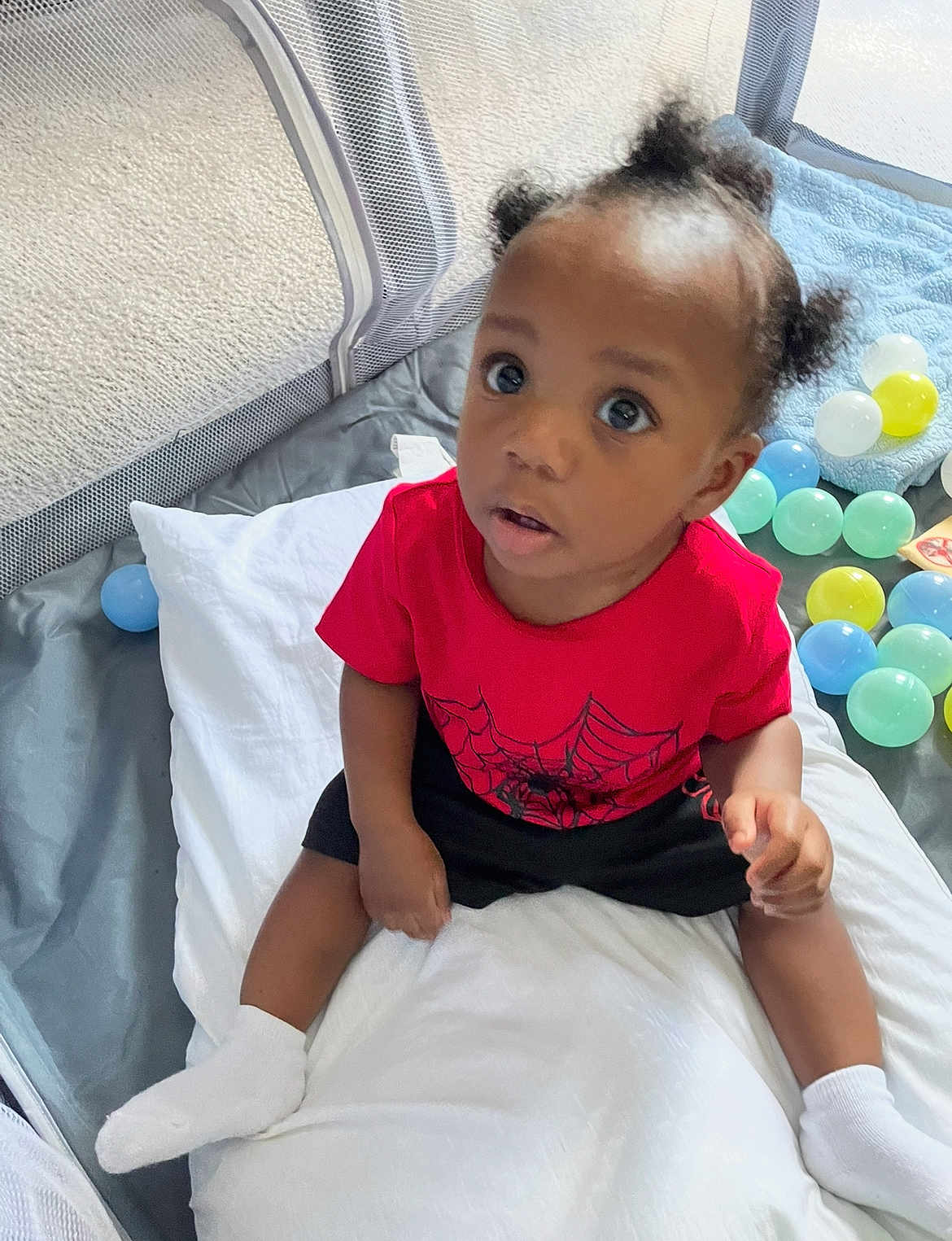 Omari is registered to the contest to win money with this photo: toddler, child, baby, pillow, playpen, plastic_balls, red_shirt, spider_web, socks, carpet, indoor, curious, sitting, person, cute, hair_puffs, toy, floor, looking_up, young_child