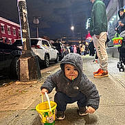 Nader is registered to the contest to win money with this photo: toddler, child, bucket, sidewalk, night, street, people, jacket, hoodie, face_paint, lollipop, cars, man, street_lamp, urban, casual_clothing, sneakers, crowd, city, outdoor