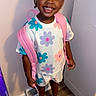 child, toddler, smiling, portrait, standing, indoor, backpack, pink_backpack, flower_pattern, colorful_clothing, braided_hair, hair_buns, earrings, shoes, clogs, hallway, door, light_switch, wood_floor, school_ready