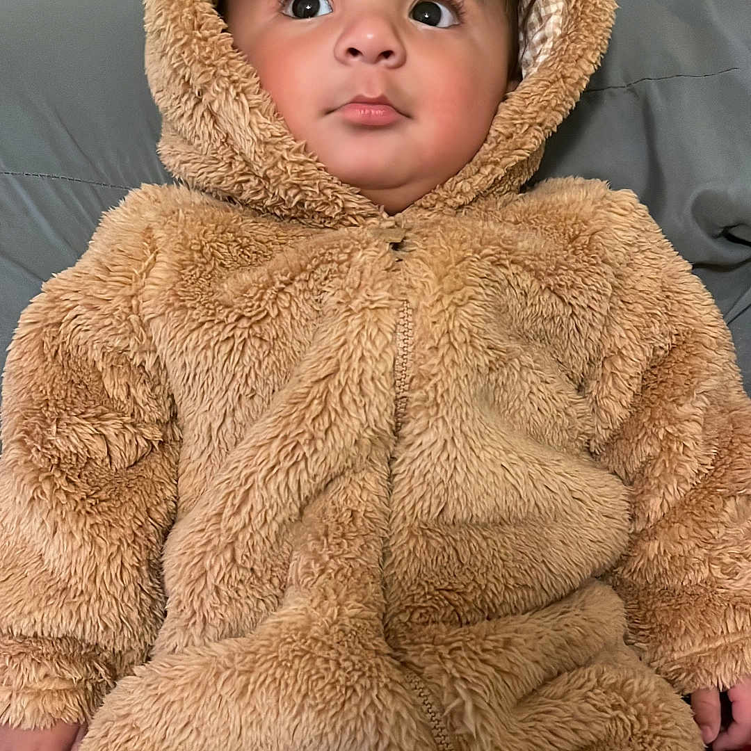 Ricardo Josiel is registered to the contest to win money with this photo: baby, bed, brown, child, cozy, cute, face, fuzzy_clothing, hands, hooded_outfit, indoors, infant, legs, looking_at_camera, pillows, portrait, sitting, soft_texture, teddy_bear_costume, wide_eyed