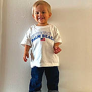 Roméo a rejoint le concours — aidez-le/la à gagner de superbes lots ! toddler, child, smiling, standing, white_tshirt, text, american_flag, jeans, rolled_jeans, sneakers, indoor, wall, plain_background, casual_clothing, portrait, happy, cute, person, footwear, short_hair