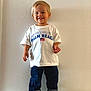 toddler, child, smiling, standing, white_tshirt, text, american_flag, jeans, rolled_jeans, sneakers, indoor, wall, plain_background, casual_clothing, portrait, happy, cute, person, footwear, short_hair