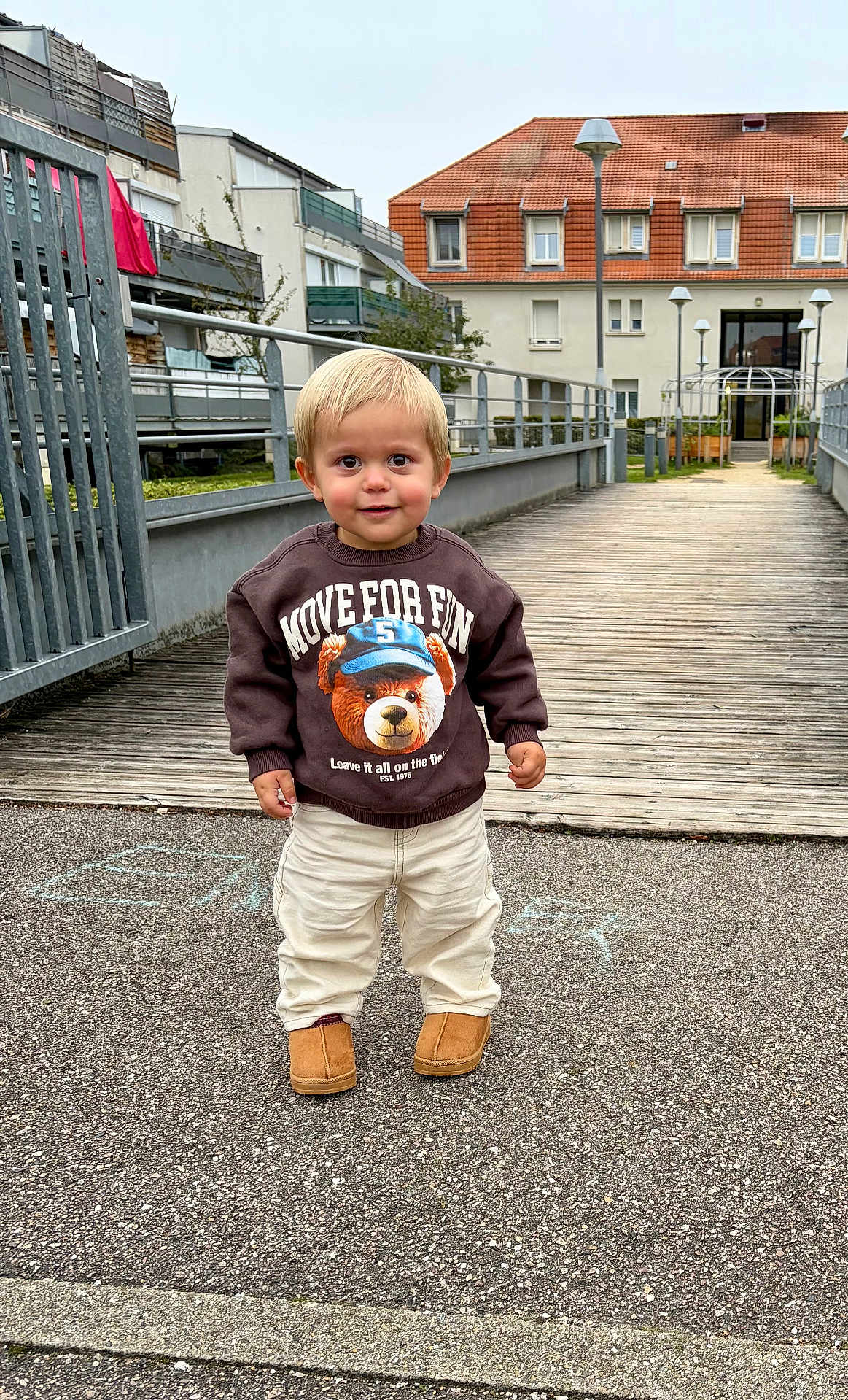 Roméo a rejoint le concours — aidez-le/la à gagner de superbes lots ! toddler, child, sweatshirt, bear_graphic, pants, boots, sidewalk, wooden_pathway, residential_buildings, fence, outdoor, daylight, person, smiling, cute, standing, urban, casual_clothing, playful, happy
