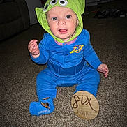 Ezra is registered to the contest to win money with this photo: baby, child, infant, costume, hat, alien_hat, blue_outfit, carpet, sign, wooden_sign, six_months, sitting, indoors, living_room, cute, smiling, person, toy_story, disney, sock