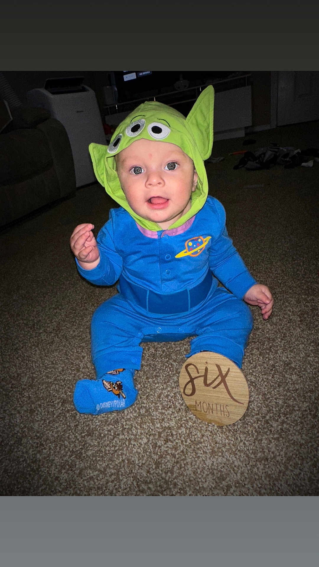 Ezra is registered to the contest to win money with this photo: baby, child, infant, costume, hat, alien_hat, blue_outfit, carpet, sign, wooden_sign, six_months, sitting, indoors, living_room, cute, smiling, person, toy_story, disney, sock