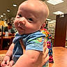 Ryder is registered to the contest to win money with this photo: baby, child, indoor, library, bookshelf, person, blue_clothing, table, hand, face, curious, seated_person, lamp, furniture, colorful_backpack, skin, cute, infant, holding, expression