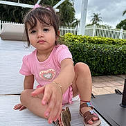 Dafne is registered to the contest to win money with this photo: toddler, child, girl, pink_outfit, sandals, outdoor, lounge_chair, white_fence, greenery, plants, curious, reaching, short_hair, bracelet, cloudy_sky, pavement, summer, daytime, portrait, cute