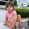 toddler, child, girl, pink_outfit, sandals, outdoor, lounge_chair, white_fence, greenery, plants, curious, reaching, short_hair, bracelet, cloudy_sky, pavement, summer, daytime, portrait, cute