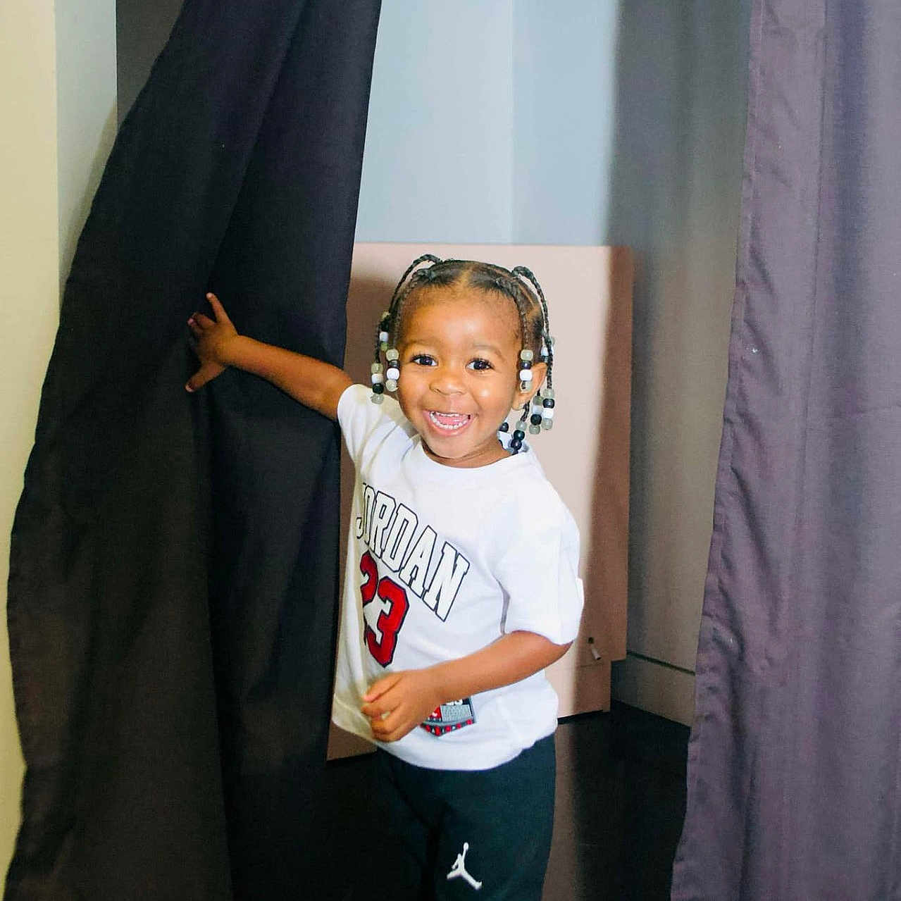 Jeremy is registered to the contest to win money with this photo: beads, black_hair, braids, casual, child, clothing, curtain, fashion, happiness, indoor, joy, pants, person, portrait, shirt, shoes, smiling, sneakers, standing, toddler