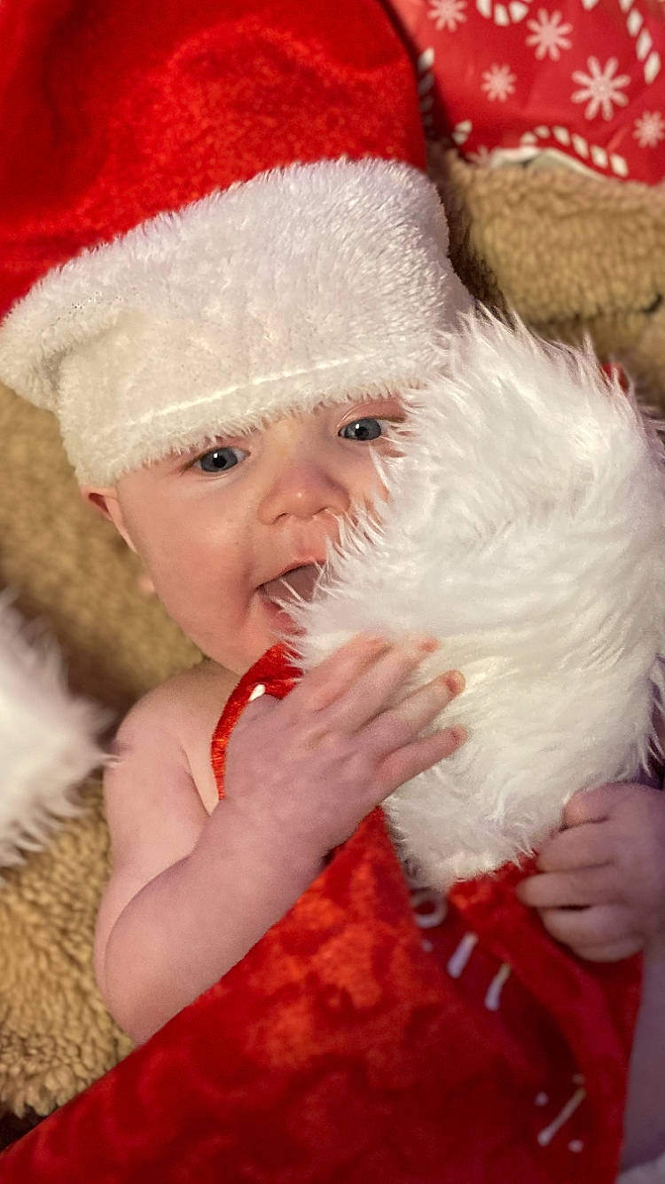 Kohen is registered to the contest to win money with this photo: baby, baby_toddler_clothing, cap, child, christmas, costume_hat, event, fictional_character, finger, fur, fur_clothing, gesture, happy, hat, head, headwear, holiday, lap, person, santa_claus
