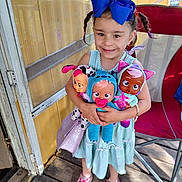 Isabella is registered to the contest to win money with this photo: child, girl, doll, blue_bow, braided_hair, dress, sneakers, porch, wooden_floor, toys, smiling, happy, playtime, pigtails, outdoor, sunlight, colorful, cute, accessory, person