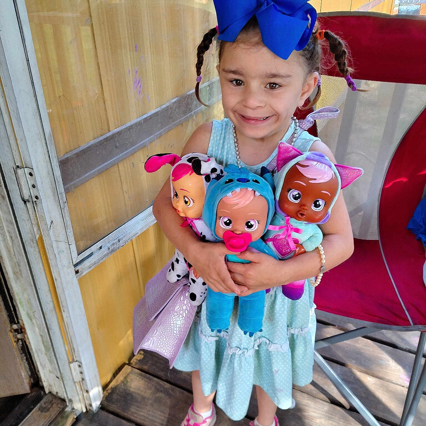 Isabella is registered to the contest to win money with this photo: child, girl, doll, blue_bow, braided_hair, dress, sneakers, porch, wooden_floor, toys, smiling, happy, playtime, pigtails, outdoor, sunlight, colorful, cute, accessory, person