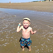 Roméo participe au concours pour gagner de l'argent avec cette photo : child, boy, beach, water, swim_shorts, cap, smiling, sunny, summer, sand, shore, playful, outdoor, vacation, happy, person, nature, daytime, recreation, fun