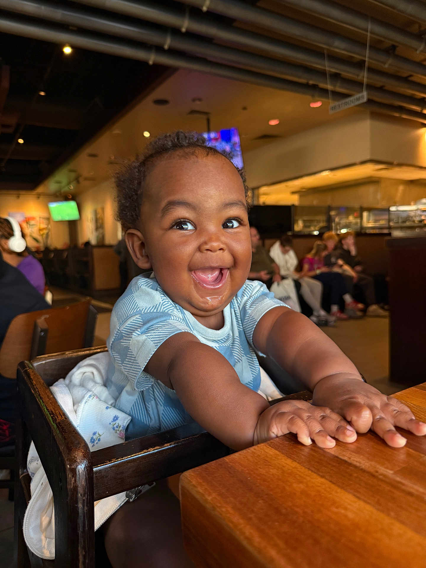 Noah joined the competition — help win amazing prizes! child, baby, toddler, high_chair, wooden_table, smile, joyful, restaurant, dining, people, face, eyes, hands, clothing, interior, ceiling_pipes, tv_screen, background, chair, infant