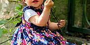 Ambre a rejoint le concours — aidez-le/la à gagner de superbes lots ! child, toddler, girl, blue_eyes, floral_dress, outdoor, nature, moss, twig, grass, leaves, stone_wall, smiling, curly_hair, playful, sitting, sunlight, shoes, greenery, portrait