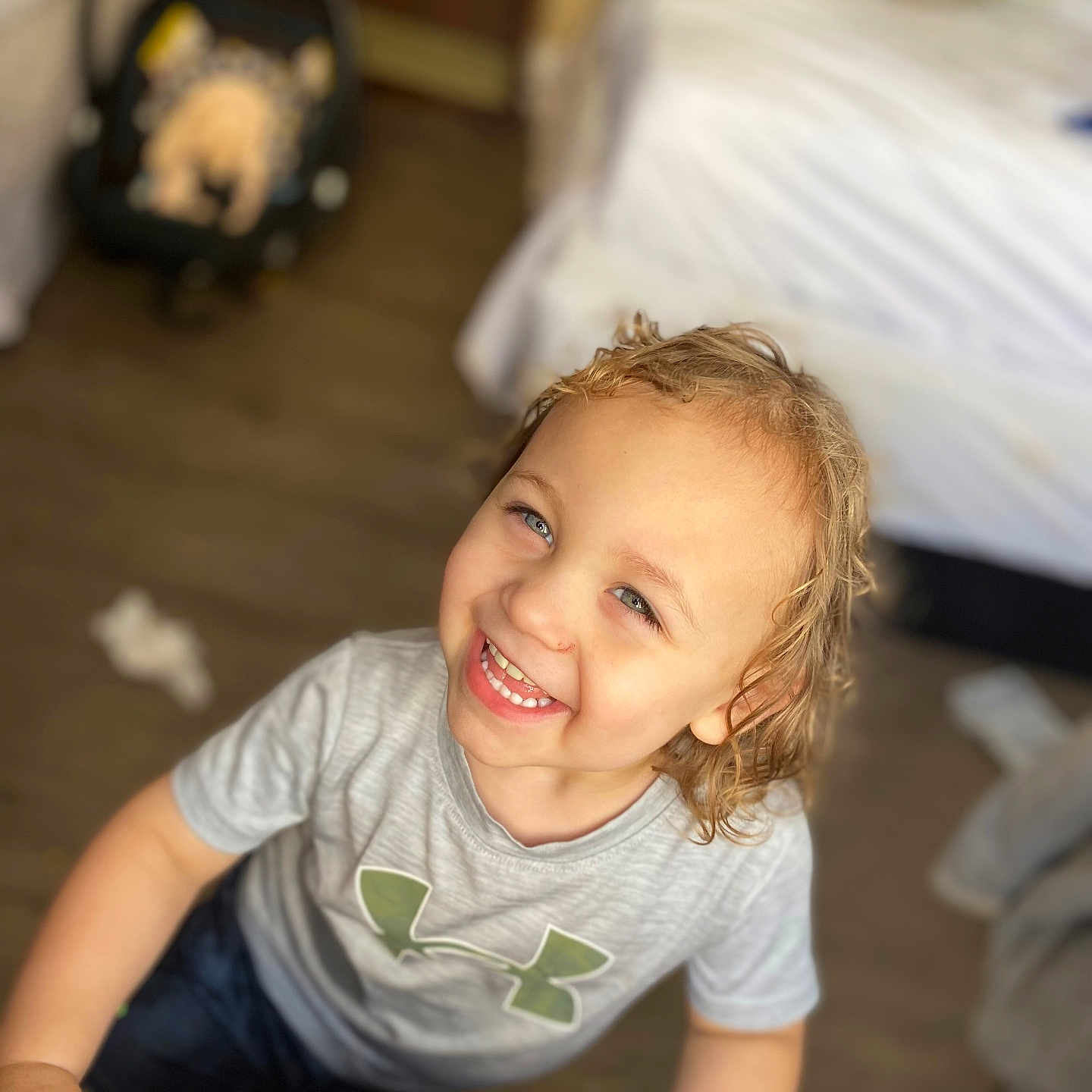 Jakavia is registered to the contest to win money with this photo: baby_car_seat, bed, blurry_background, casual_clothing, cheerful, child, curly_hair, floor, green_logo, grey_shirt, happy, indoor, light_skin, looking_up, person, portrait, short_sleeves, smiling, toddler, wood_floor