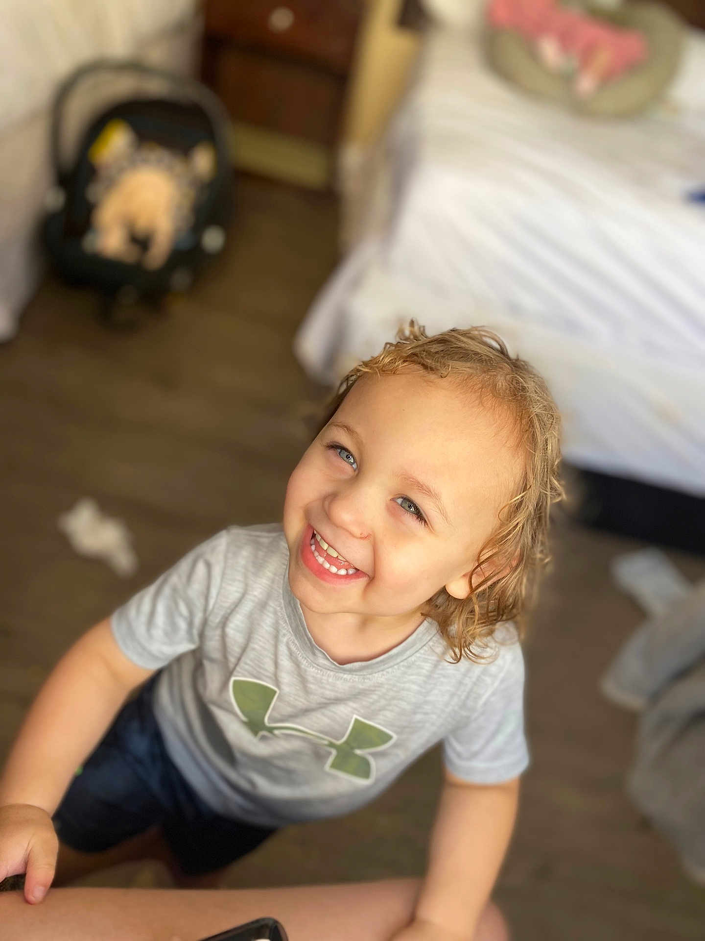 Jakavia is registered to the contest to win money with this photo: child, smiling, indoor, curly_hair, grey_shirt, green_logo, bed, baby_car_seat, floor, happy, person, portrait, looking_up, casual_clothing, wood_floor, blurry_background, light_skin, short_sleeves, toddler, cheerful