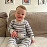 baby, child, smiling, striped_pajamas, couch, beige, indoor, family_photos, wall, framed_photos, happy, portrait, sitting, feet, furniture, infant, cute, home, person, cozy