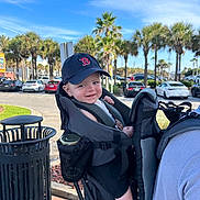 Cade is registered to the contest to win money with this photo: baby, child, hat, cap, smile, face, person, baby_carrier, outdoor, daylight, sky, clouds, palm_trees, parking_lot, cars, trash_can, clothing, shoulder, backpack, happy