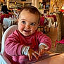toddler, child, high_chair, pink_sweater, menu, restaurant, indoor, smiling, happy, blue_eyes, table, chair, lighting, decor, person, cute, baby, face, clothing, furniture