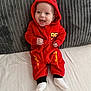 Cody is registered to the contest to win money with this photo: baby, child, red_onesie, hood, smiling, sitting, blanket, couch, cushion, happy, infant, clothing, portrait, indoor, cute, person, footwear, white_socks, cozy, young