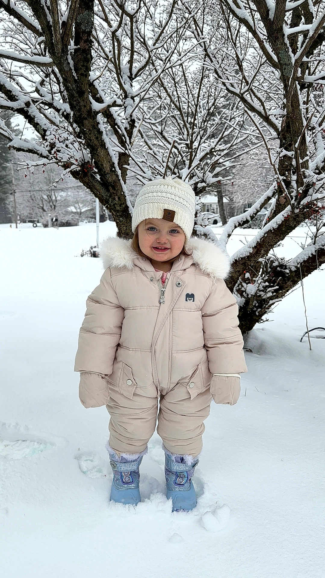 Sophie is registered to the contest to win money with this photo: toddler, child, snow, winter_clothing, snowsuit, knit_hat, boots, outdoor, snowy_tree, winter, smiling, cold_weather, nature, park, daylight, cute, portrait, standing, season, frost