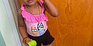Alani is registered to the contest to win money with this photo: child, girl, indoor, standing, beads, colorful_hair, pink_top, black_shorts, black_shoes, toy, wooden_door, floor_tiles, bracelet, necklace, curly_hair, expression, person, casual_clothing, young_child, accessories
