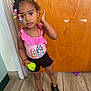 child, girl, indoor, standing, beads, colorful_hair, pink_top, black_shorts, black_shoes, toy, wooden_door, floor_tiles, bracelet, necklace, curly_hair, expression, person, casual_clothing, young_child, accessories