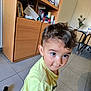 toddler, child, curly_hair, yellow_shirt, indoor, floor_tiles, cabinet, furniture, curious, big_eyes, person, young_child, home, casual_clothing, brown_hair, hair, standing, room, household_items, light