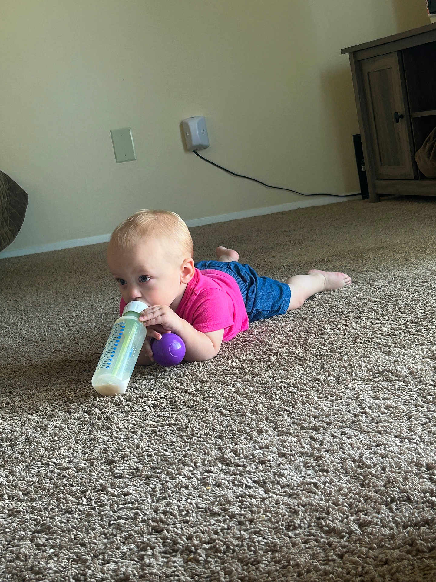 Sophie is registered to the contest to win money with this photo: toddler, baby, carpet, floor, bottle, toy, purple, pink_shirt, blue_shorts, indoor, child, person, crawling, curious, home, cabinet, wall, power_outlet, foot, head