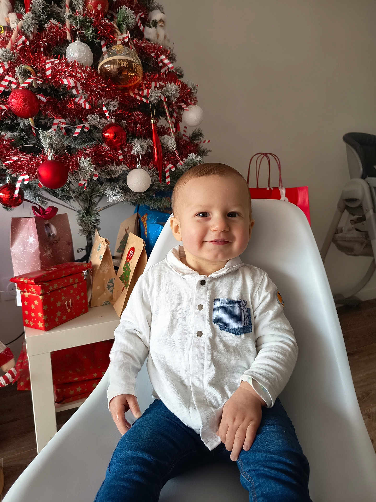 Manolo a rejoint le concours — aidez-le/la à gagner de superbes lots ! toddler, child, smile, white_shirt, blue_jeans, chair, christmas_tree, ornaments, gifts, holiday, indoor, decorations, festive, red, white, present, table, happy, person, celebration
