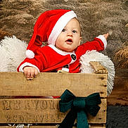 Manolo a rejoint le concours — aidez-le/la à gagner de superbes lots ! baby, child, santa_hat, christmas_costume, wooden_crate, green_bow, fluffy_pillow, winter_background, festive, holiday, portrait, cute, red_clothing, indoor, smiling, person, infant, cozy, decor, seasonal