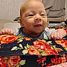 baby, infant, child, portrait, smiling, happy, face, eyes, hands, indoor, blanket, floral_pattern, clothing, gray_shirt, bedroom, furniture, cozy, close_up, sitting, photography