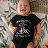 baby, bed, black_outfit, blanket, child, clothing, cozy, cute, face, floral_blanket, grinning, happy, indoor, infant, lying_down, onesie, photography, portrait, skeleton_graphic, smiling