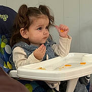 Jade is registered to the contest to win money with this photo: baby, blue, child, clothing, cute, eating, face, focused, food, high_chair, indoor, person, pigtails, polka_dot, seat, snack, table, toddler, white, young_child