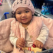 Amerie is registered to the contest to win money with this photo: aisle, baby, child, clothing, curly_hair, cute, face, hand, happy, indoor, infant, jingle_bells, knit_hat, person, pink_vest, shopping_cart, sitting, smile, store, toy