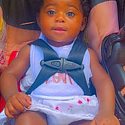 Faith is registered to the contest to win money with this photo: baby, child, car_seat, safety_harness, white_dress, floral_embroidery, curly_hair, brown_skin, infant, cute, portrait, seatbelt, hands, legs, indoor, closeup, adorable, young_child, sitting, looking_at_camera