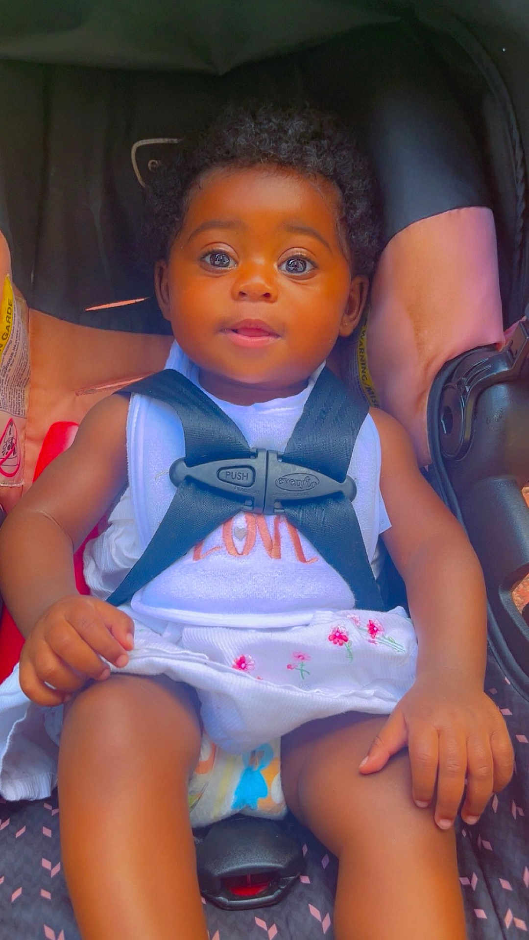 Faith is registered to the contest to win money with this photo: baby, child, car_seat, safety_harness, white_dress, floral_embroidery, curly_hair, brown_skin, infant, cute, portrait, seatbelt, hands, legs, indoor, closeup, adorable, young_child, sitting, looking_at_camera