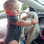 Easton is registered to the contest to win money with this photo: baby, child, car, steering_wheel, hands, blue_eyes, onesie, interior, vehicle, window, side_mirror, seat, person, cute, curious, infant, driving, dashboard, beads, hand