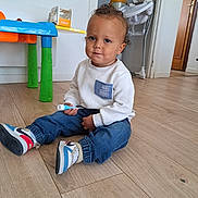 Milow participe au concours pour gagner de l'argent avec cette photo : baby, blue_jeans, casual_clothing, child, curly_hair, door, floor, highchair, holding_object, home, indoor, person, sitting, sneakers, toddler, toy, wall, white_sweater, wooden_floor, young_child