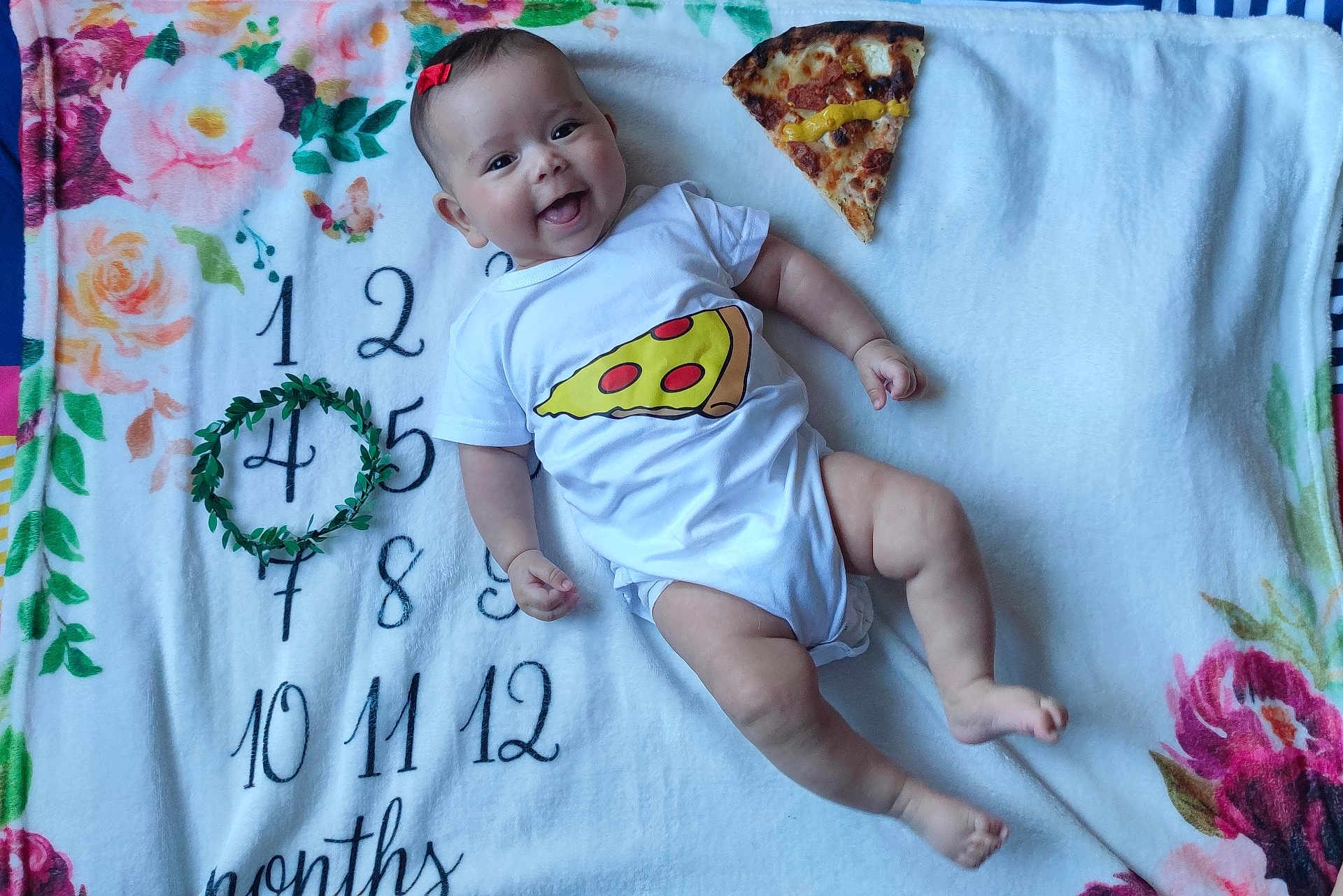 Anya participe au concours pour gagner de l'argent avec cette photo : baby, infant, smiling, onesie, pizza_slice, blanket, milestone_blanket, months, flower_pattern, red_bow, cute, happy, lying_down, indoors, child, face, skin, hand, foot, playful