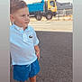 toddler, child, white_polo, blue_shorts, white_shoes, asphalt, outdoor, construction_truck, vehicle, short_hair, side_view, casual_clothing, sunny_day, standing, young_boy, curious_expression, fence, industrial_area, daylight, footwear
