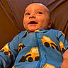 baby, infant, child, smiling, happy, portrait, close_up, face, eyes, open_mouth, indoors, seated, blue_outfit, patterned_clothing, yellow_designs, brown_background, cushion, cozy, looking_up, photography