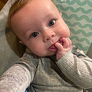 Charlie is registered to the contest to win money with this photo: baby, infant, close_up, selfie, face, hand, fingers, blue_eyes, chubby_cheeks, gray_clothing, patterned_background, curious_expression, indoors, cute, child, portrait, sitting, young, person, soft_lighting